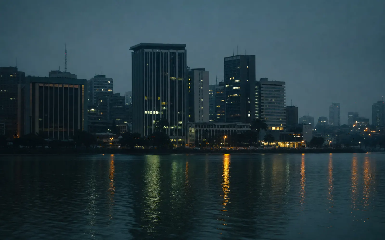 Le Plateau d'Abidjan à l'heure bleue, vu depuis la lagune : les tours allumées se reflètent sur l'eau sombre.